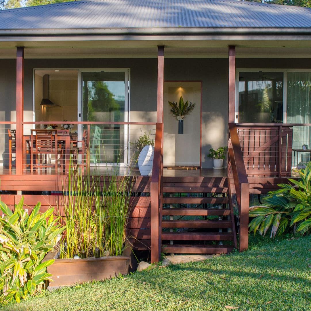 The evening sun shines on a cottage with a covered wood deck, sliding glass doors, a manicured lawn and tropical plants.