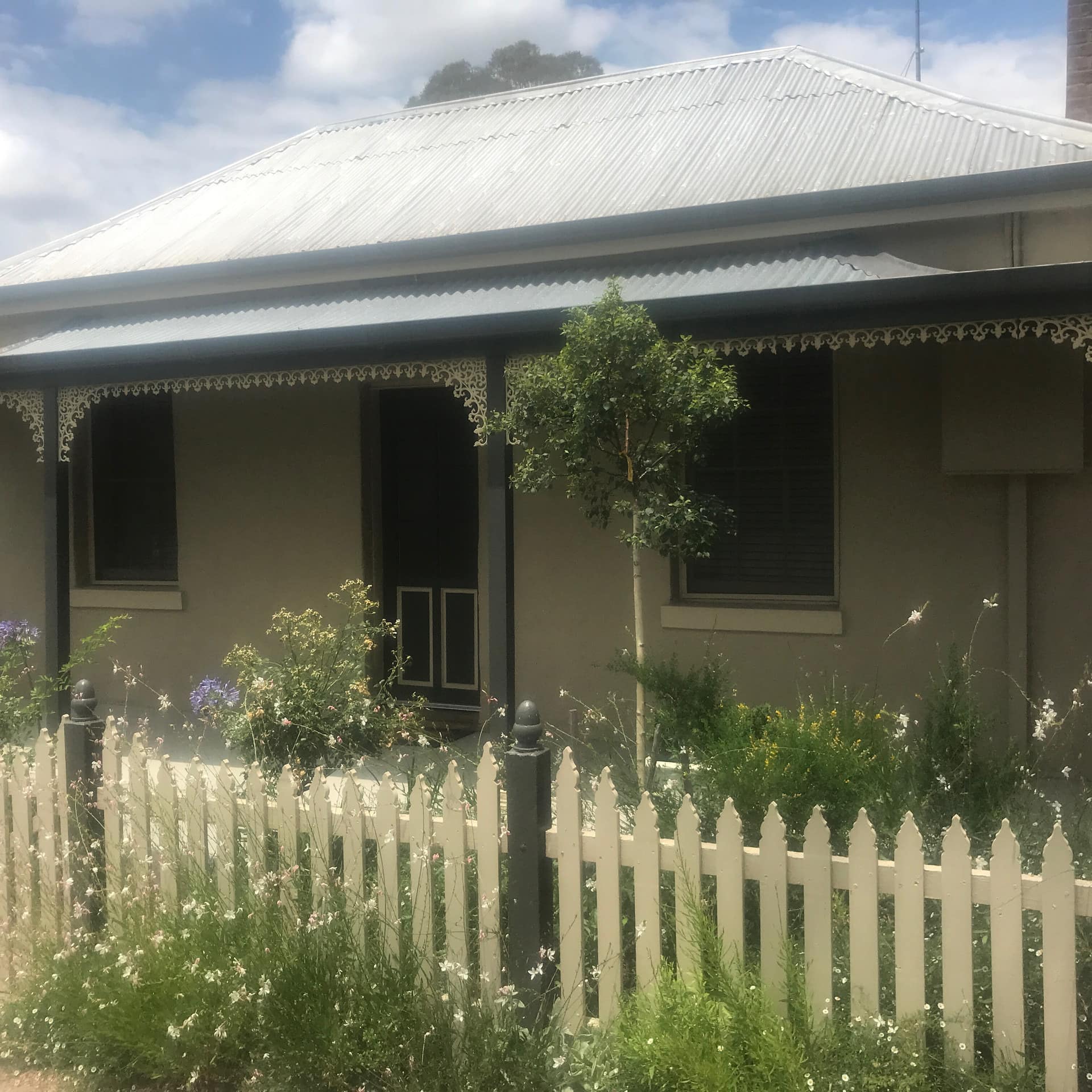 Exterior of romantic Birch Cottage with Victorian trim, a front porch, a picket fence with flowers and a tin roof.
