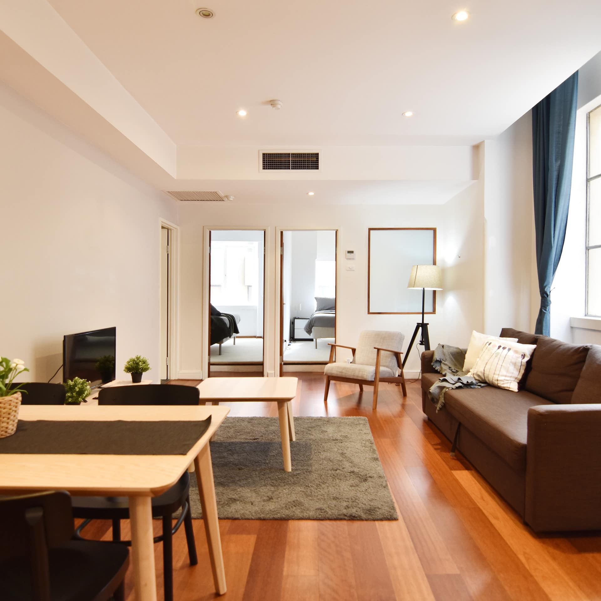 Open living room with hardwood floors, brown sofa, wooden dining set, and a wide window with blue curtains.