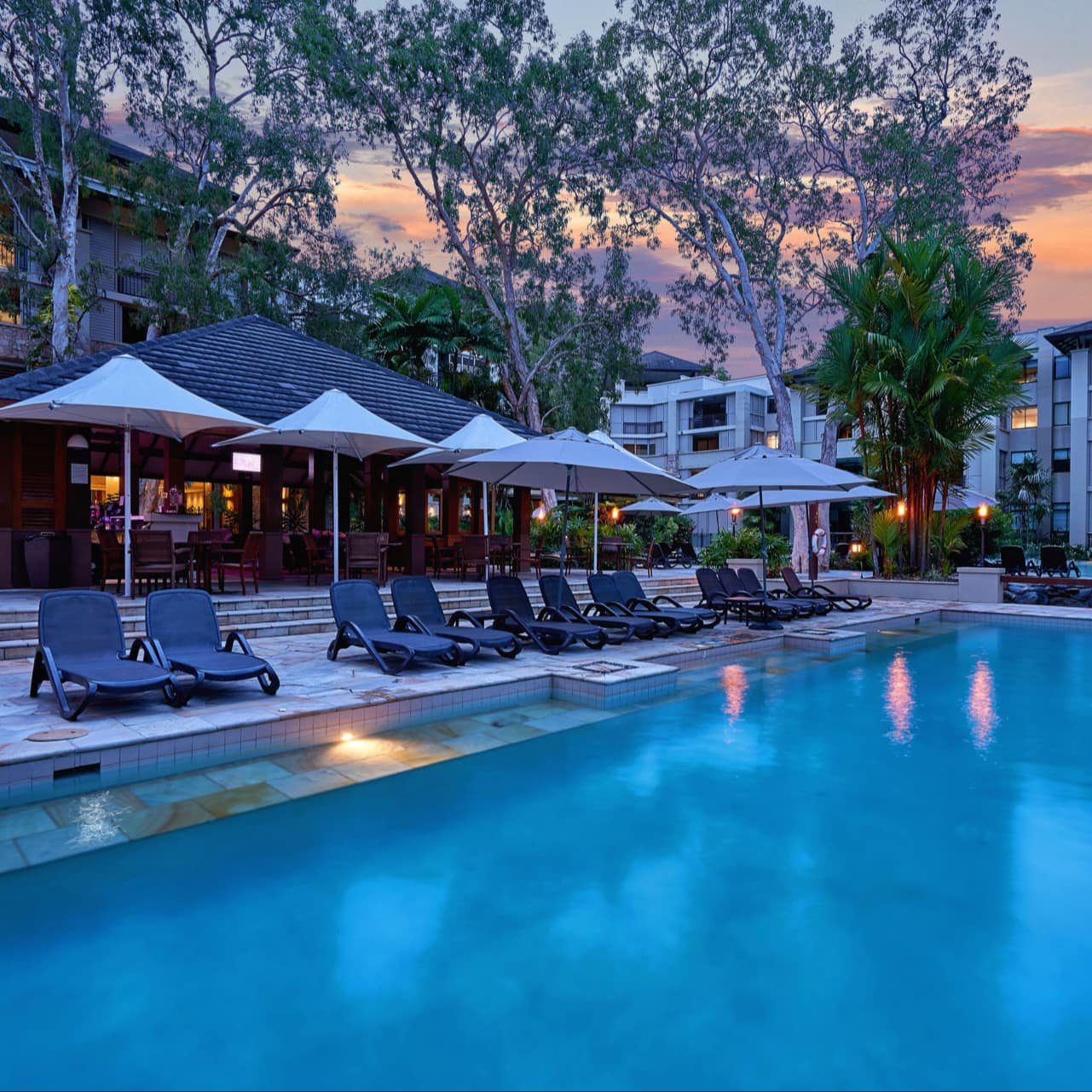 Evening view of large pool at an apartment complex with natural stone patio space and black lounge chairs.