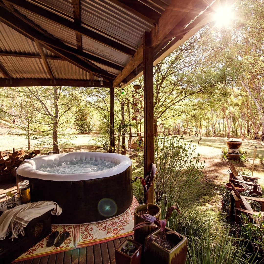 Sunlight filters through trees surrounding a covered wood patio with a hot tub, outdoor seating and tiki torches.
