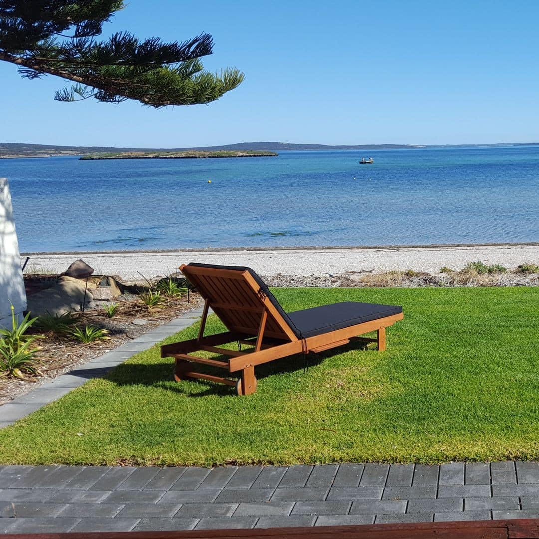 A black canvas lounge chair sits on a manicured lawn in front of a sandy beach and crystal blue water.