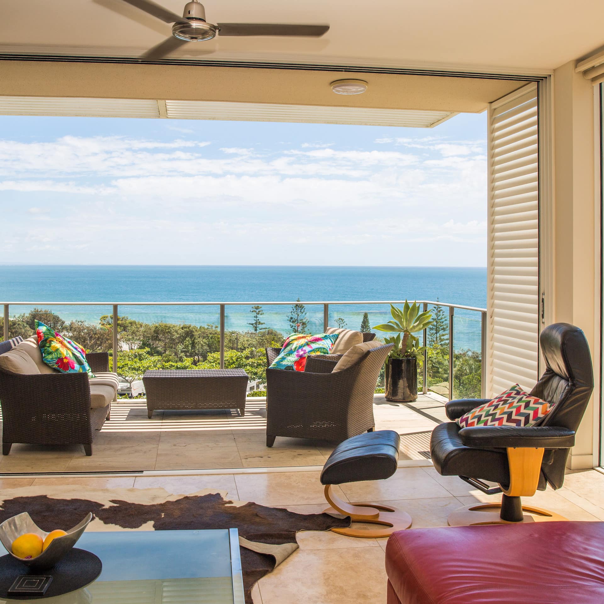 A living room with beige tile floors, leather sofa and chair, cowhide rug, open glass doors to balcony with ocean views.