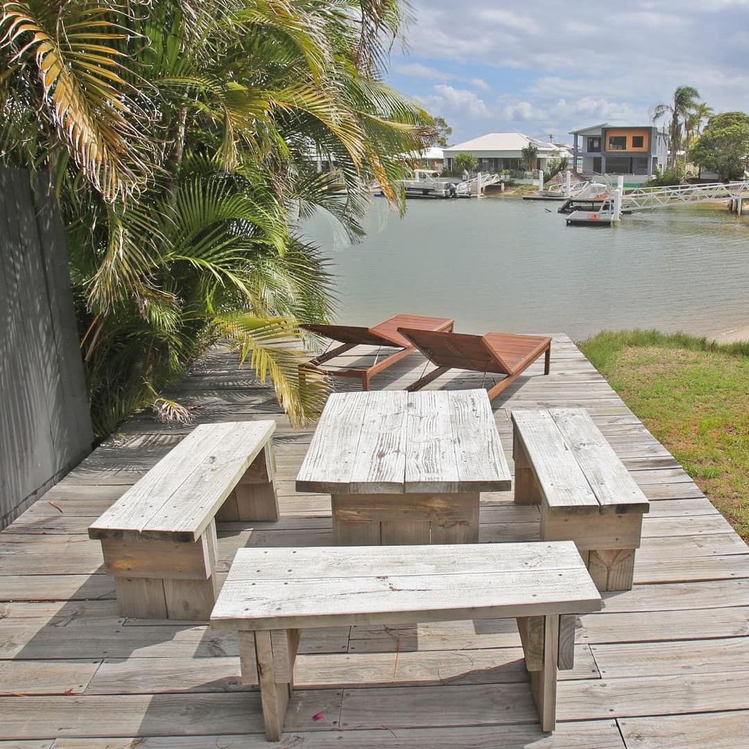 A wooden deck with a wooden table and benches, wooden lounge chairs, and palm trees, overlooking with views of the marina.