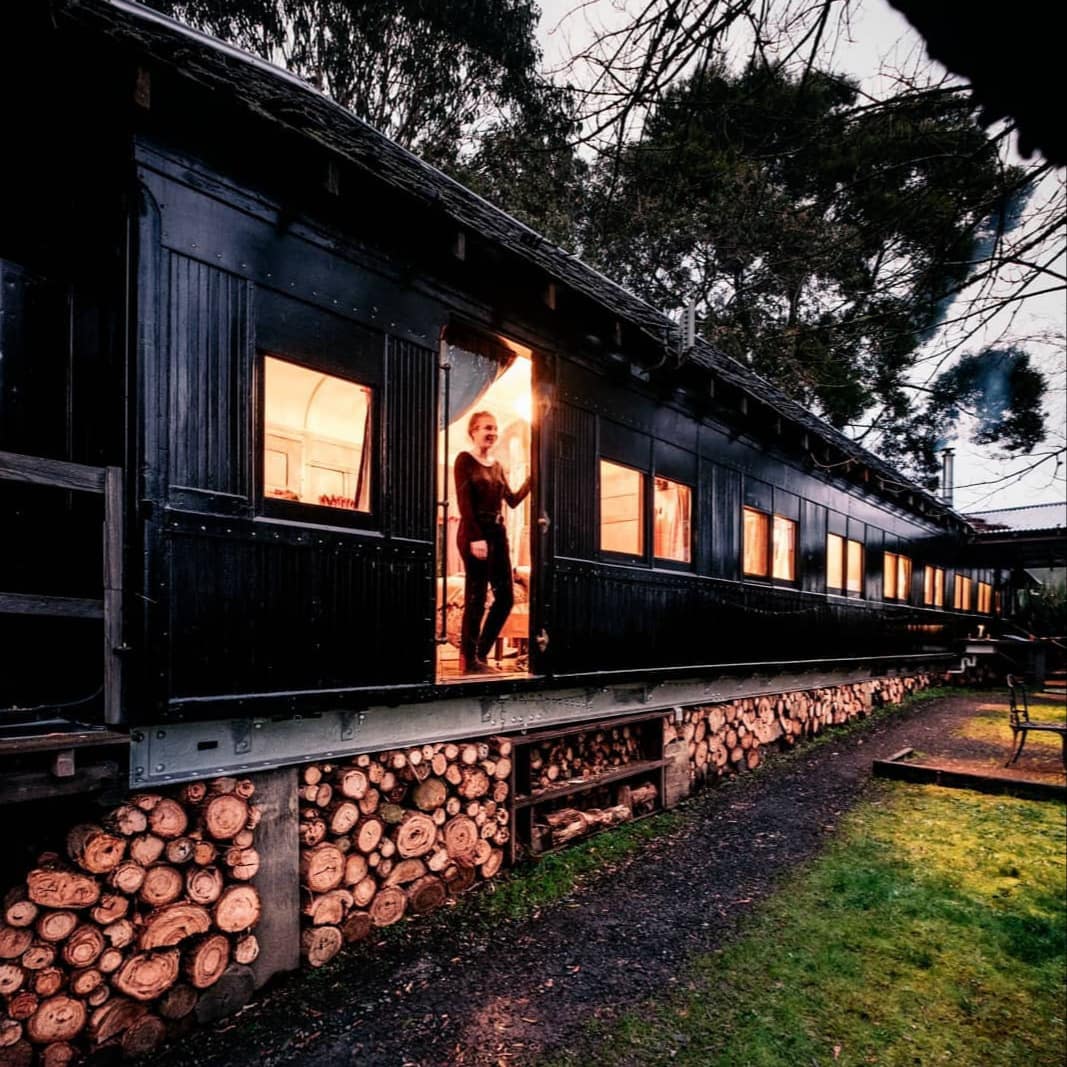 Woman stands in door of black, Victorian steam carriage converted to a Lorne cabin with firewood stacked underneath