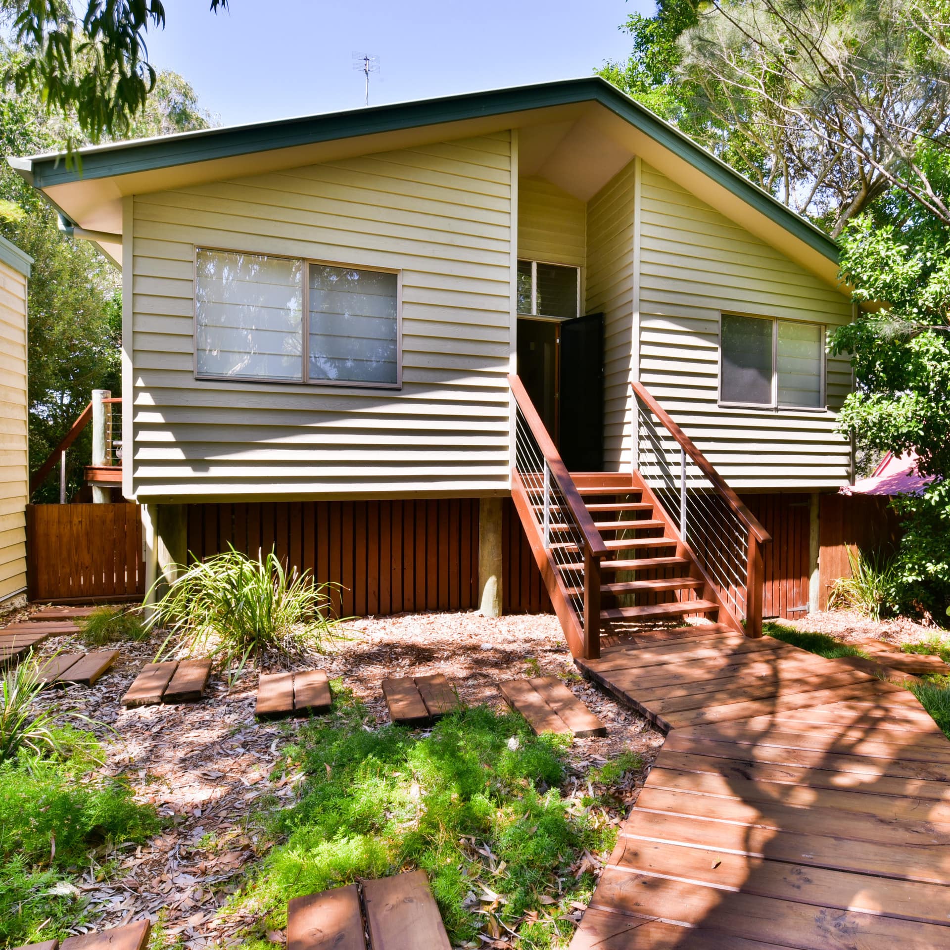Exterior view of home with yellow vinyl siding, green roof, raised wooden walkway, and lush vegetation surrounding the home.