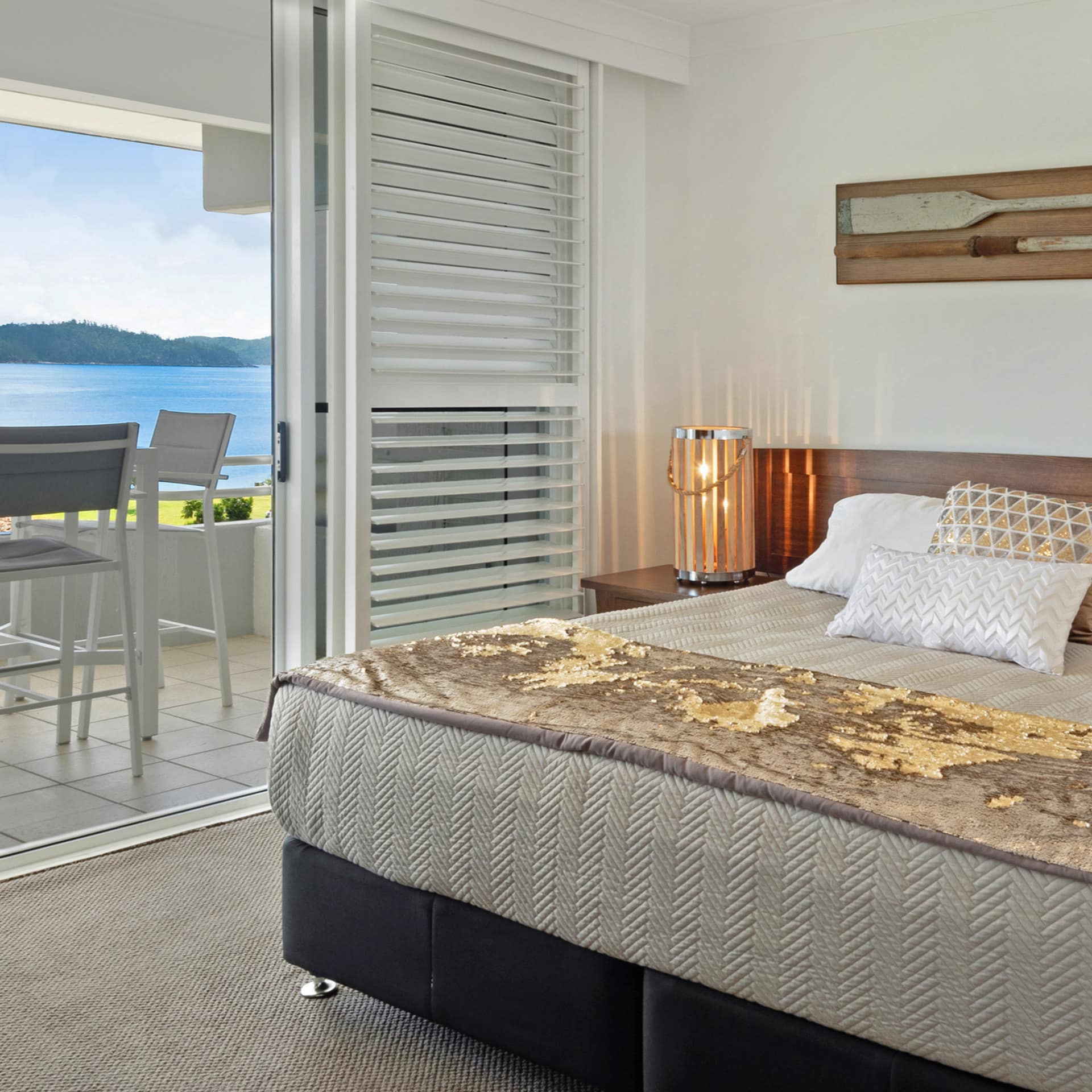 A bedroom with a king size bed, tan and gold bedding, sliding glass doors open onto attached veranda with ocean views.