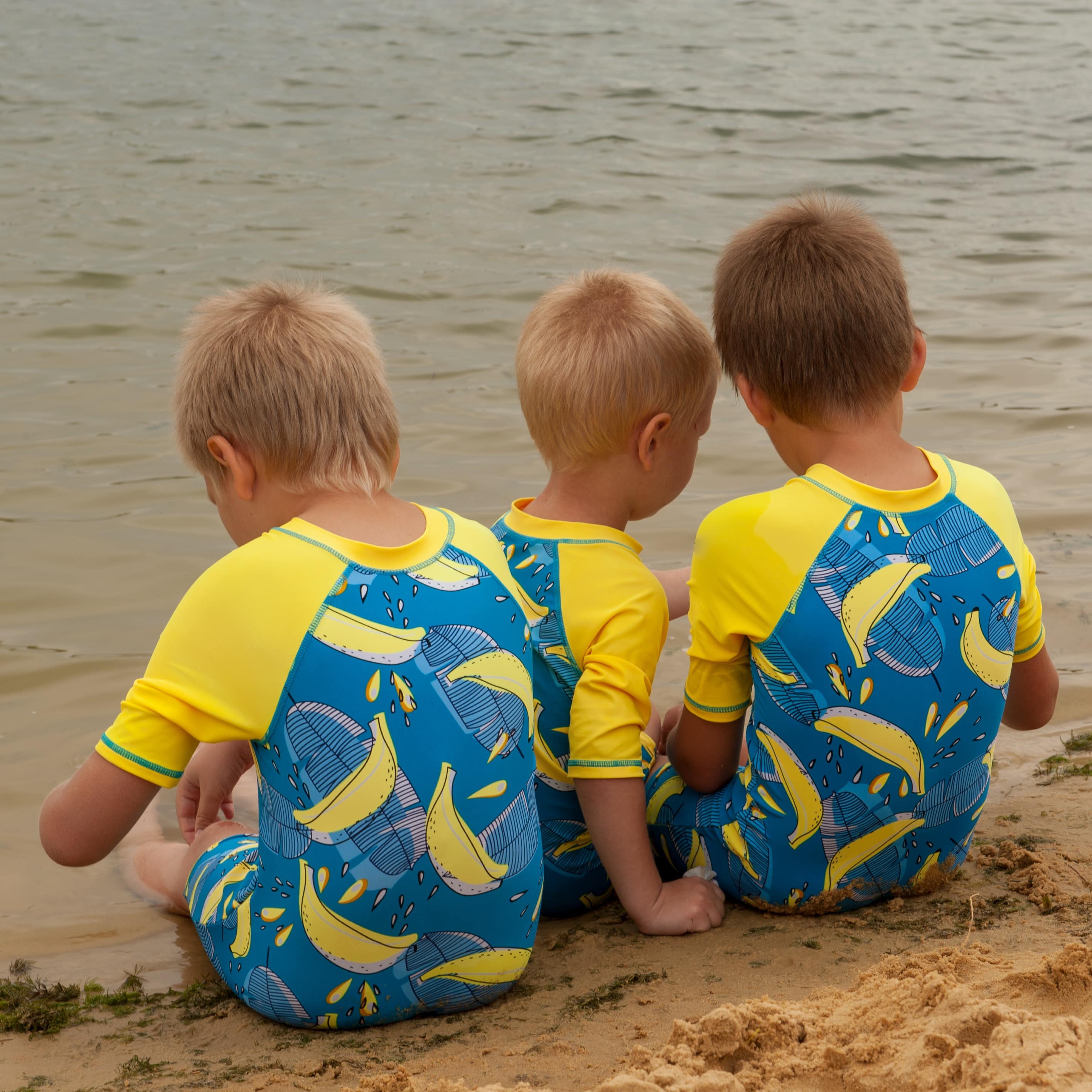 Three young boys dressed in matching swimwear sitting in sand by the river playing with their backs to the camera