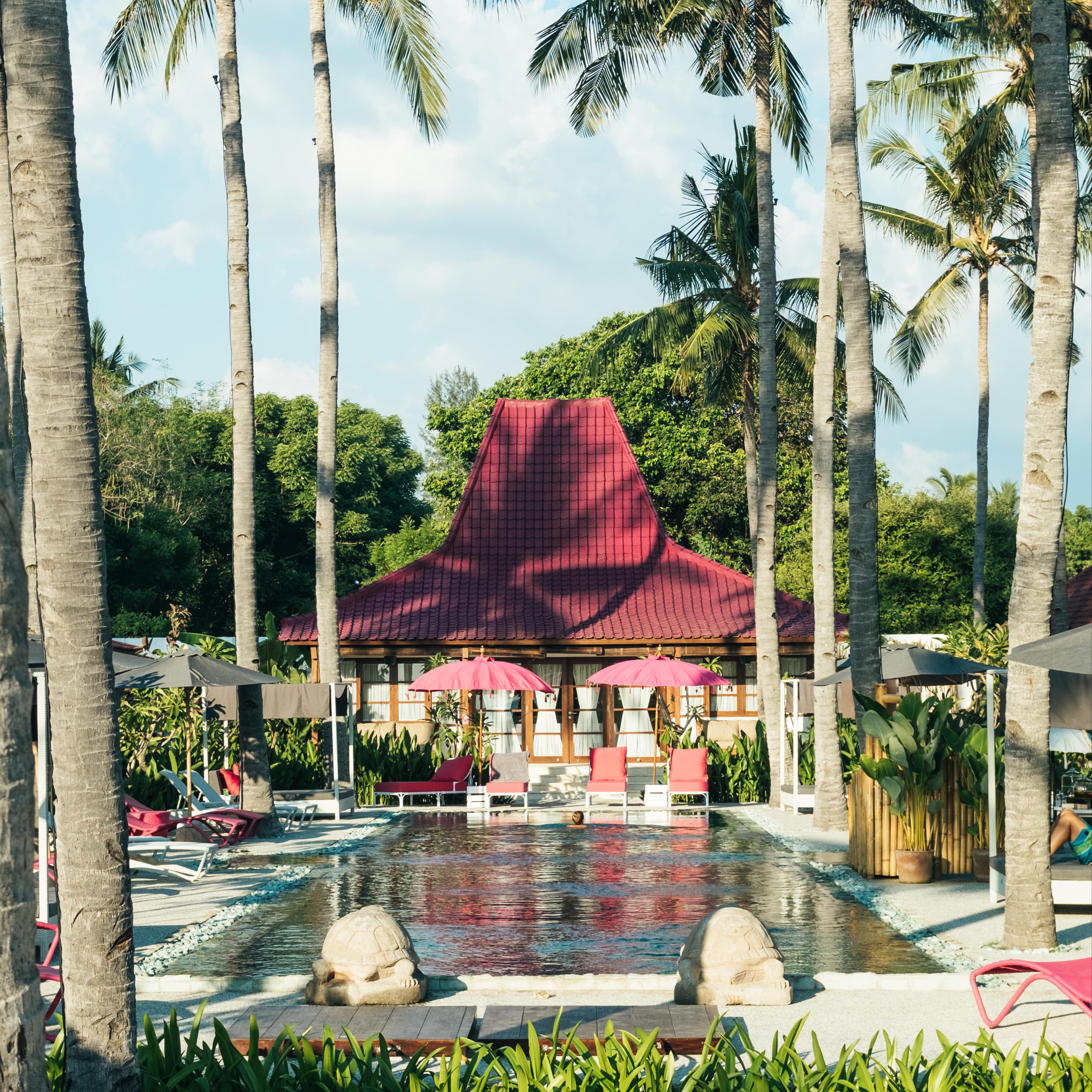 A villa with a red tile roof, many windows, a rectangular pool, turtle statues and a lounge area with chairs and umbrellas