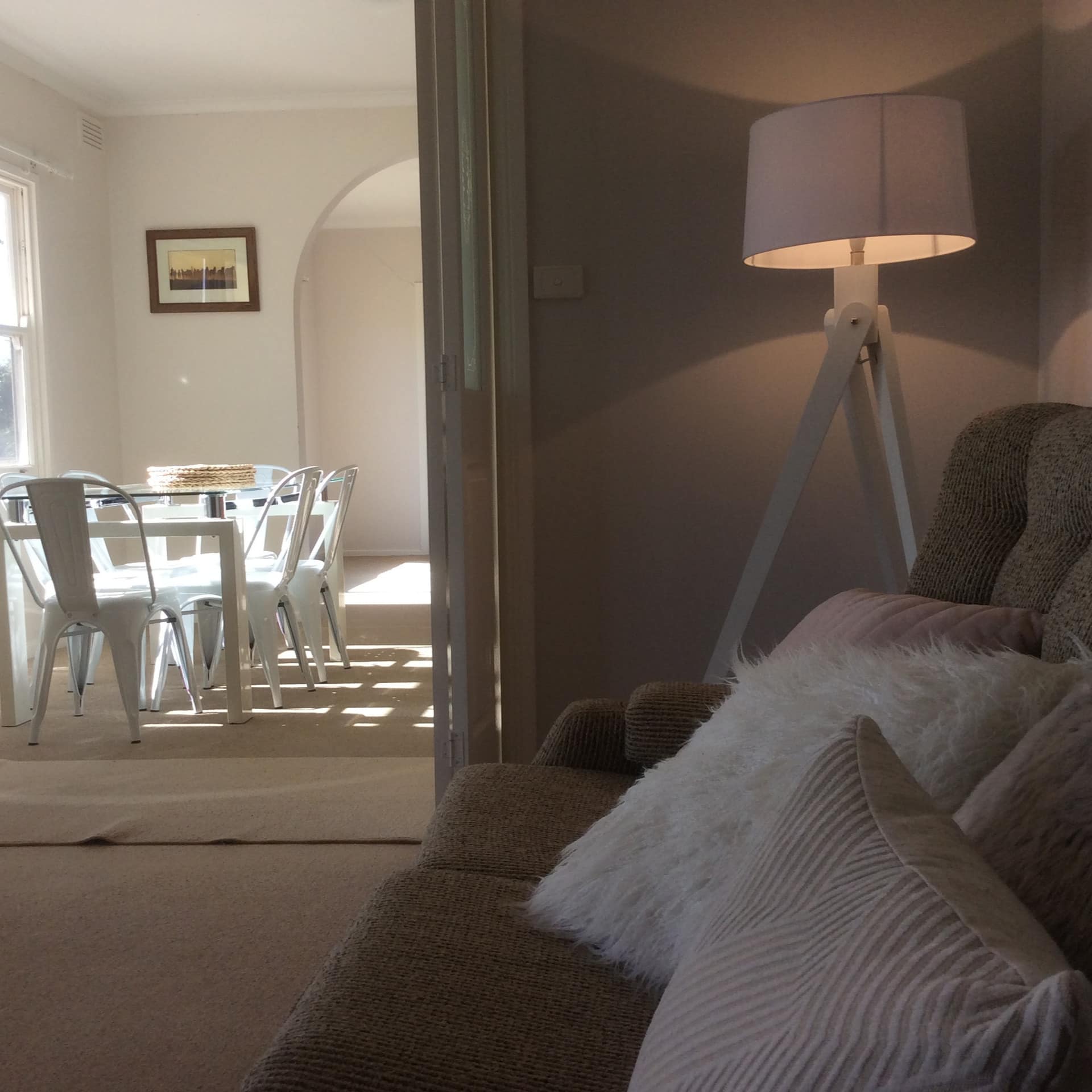 View of living and dining room, furnished in neutral tones, tan sofa, light pink pillows, glass dining table, white chairs.