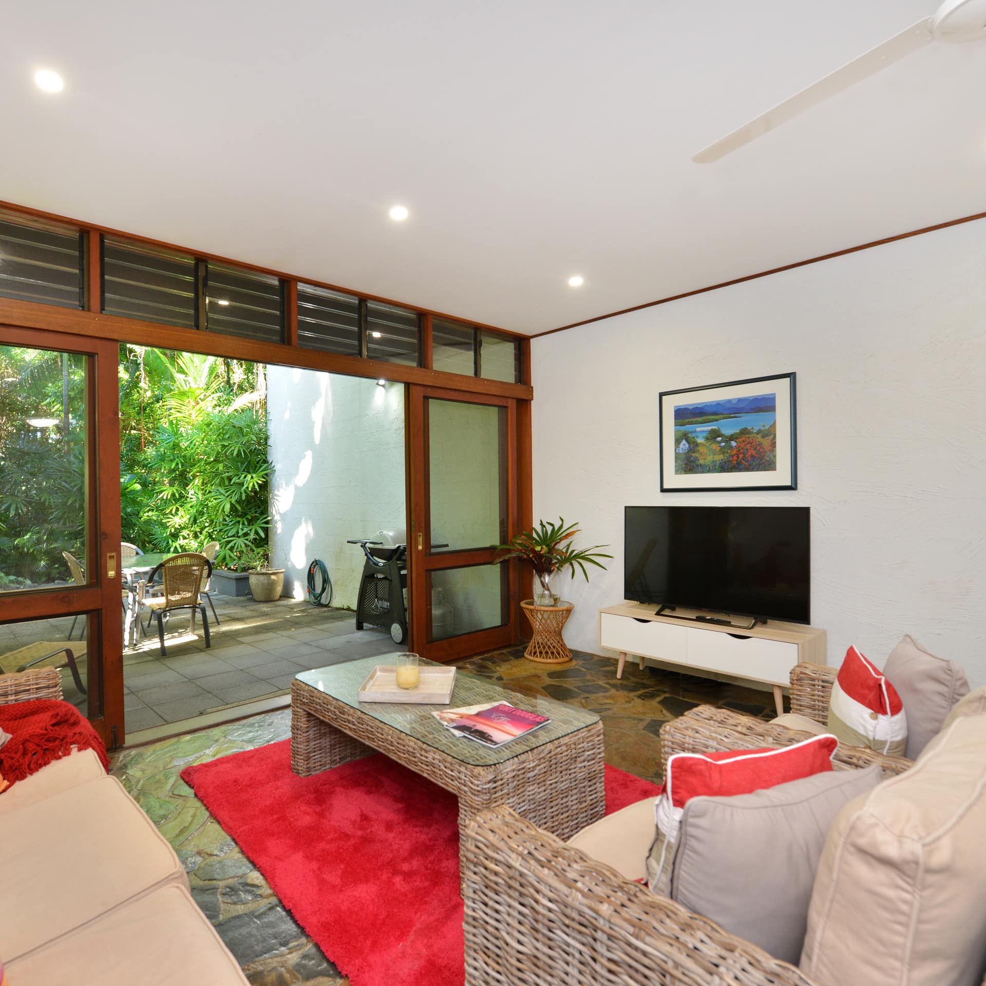 Living room with natural stone floors, beige sofas, red and orange accents, wide sliding glass doors open onto lush garden.