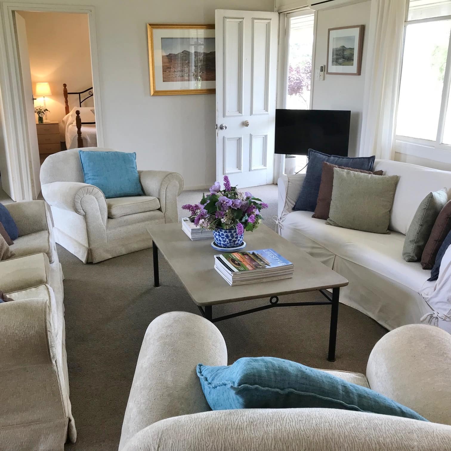 A spacious sitting room with overstuffed sofas and chairs, plush carpet, large, sunny windows and two bedrooms in background.