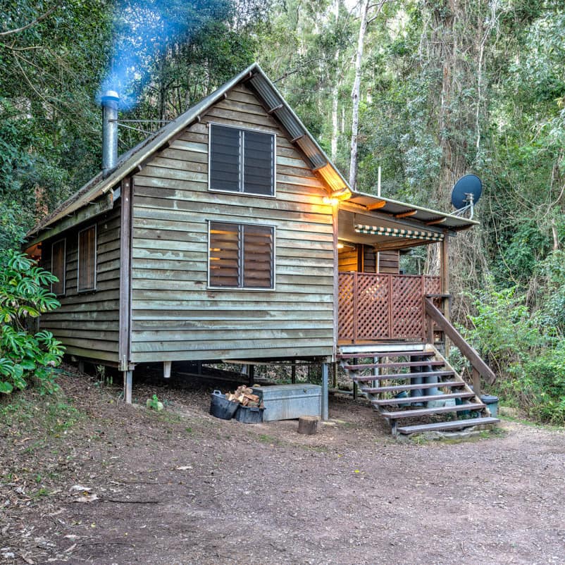 Exterior of rustic wood cabin surrounded by thick forest, stairs lead to a covered porch, smoke billows out of the chimney.