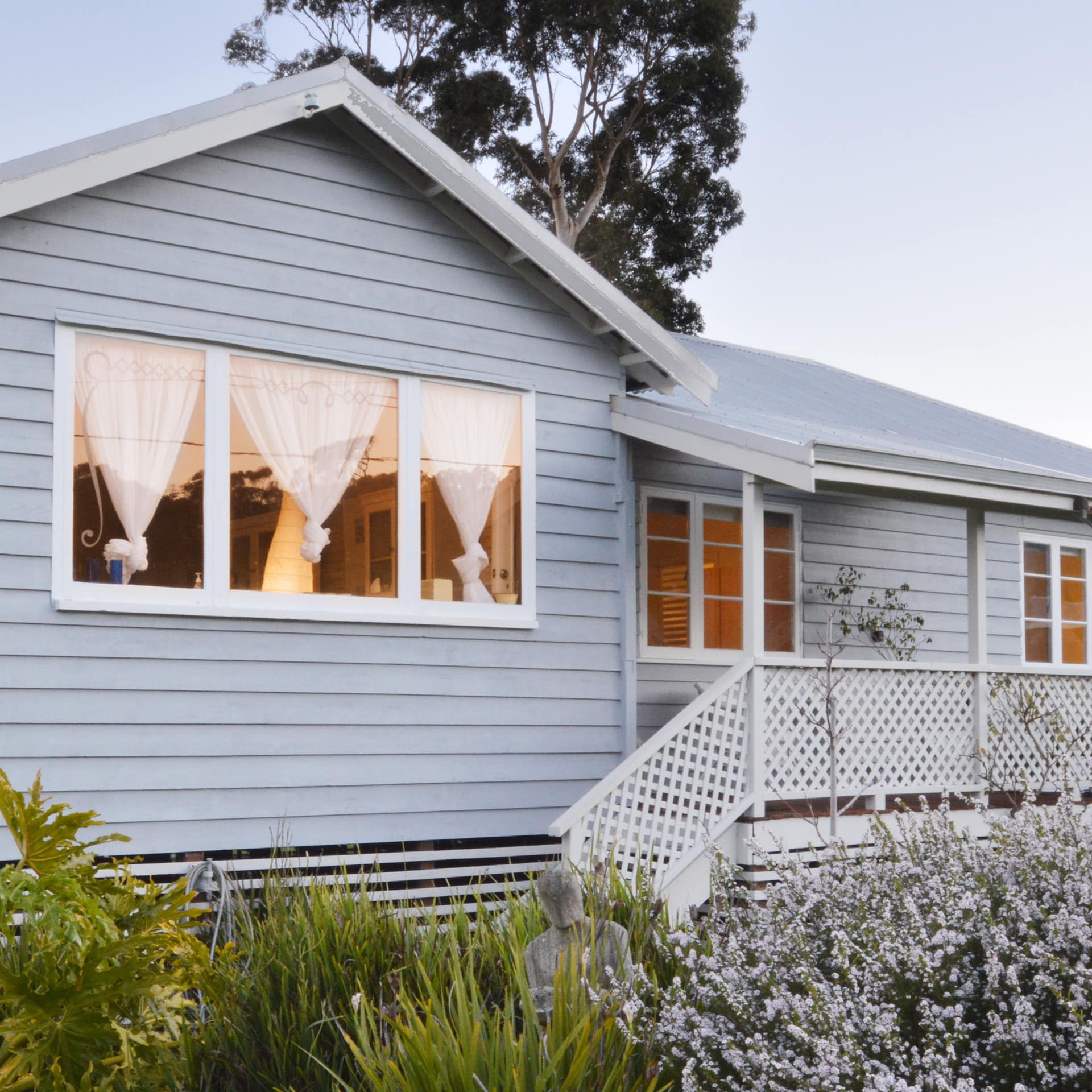 Exterior of a charming cottage with light blue siding, a covered front porch, wide windows, and a lush garden.