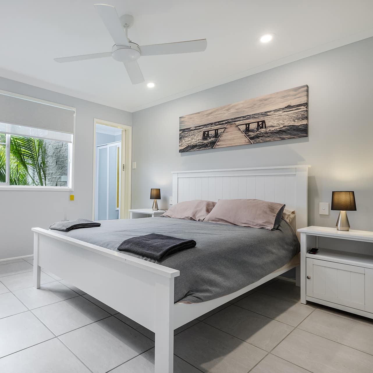 Bedroom with white tile floors, light grey walls, a king size bed, grey bedding, and a wide window with nature views.